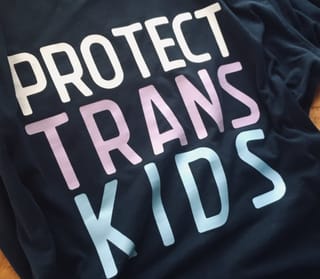 a black t-shirt that reads protect trans kids. the lettering is in the colors of the trans flag- white, pink, blue