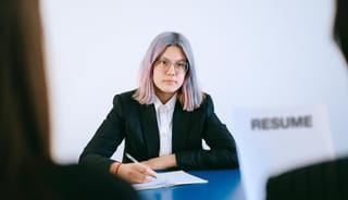 two interviewers, obscured, interview a woman with multicolor and mostly blue or pink or silver hair while holding a paper that says "resume"